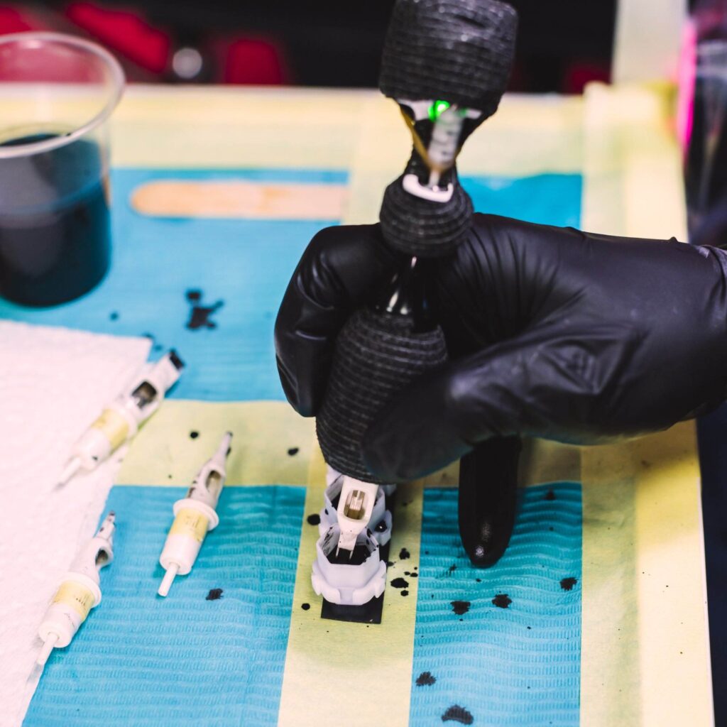 A gloved hand holds a tattoo machine above a surface covered with protective plastic, surrounded by tattoo needles and a cup of black ink.