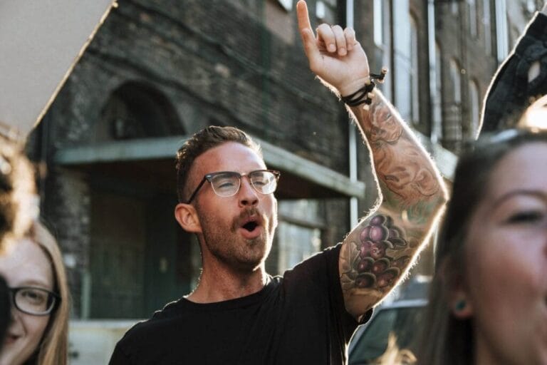 Man with glasses raising his hand, singing or shouting outdoors during daylight.