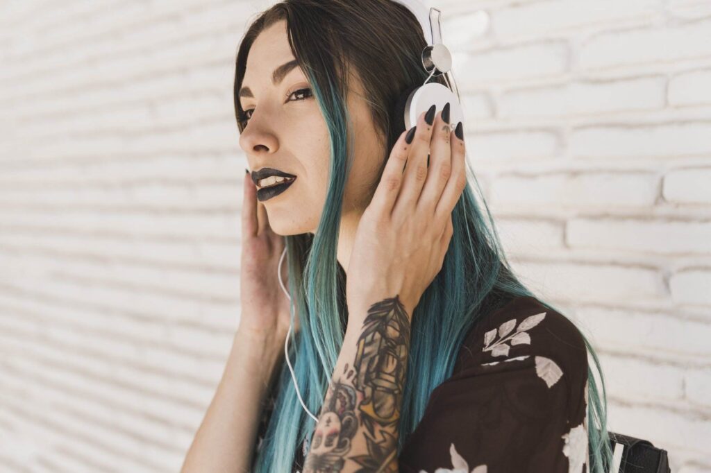 Person with long turquoise hair and tattooed arm listening to music on white headphones, standing against a white brick wall.