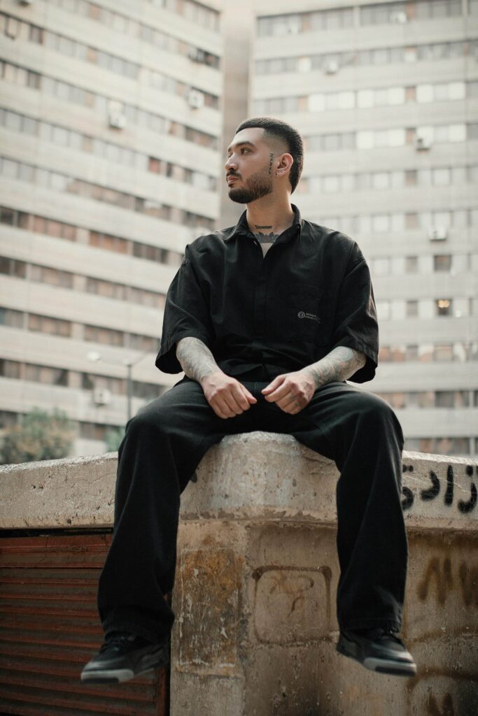 A person with tattoos sits on a concrete ledge outdoors, looking into the distance, with high-rise buildings in the background.