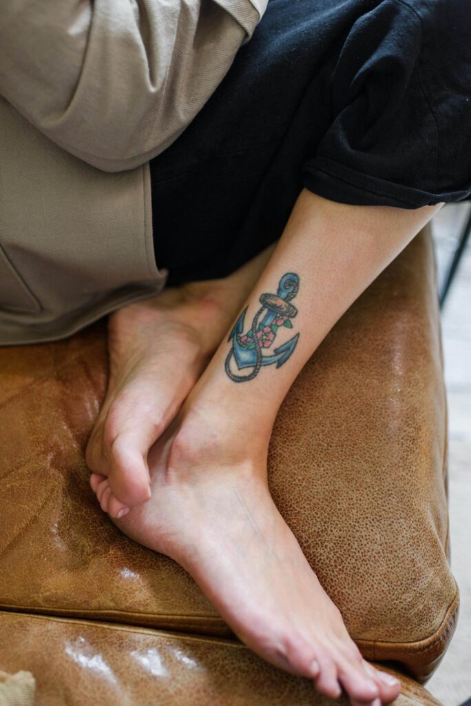 A person's lower leg with an anchor tattoo featuring red flowers and a pink ribbon, resting on a brown leather seat.