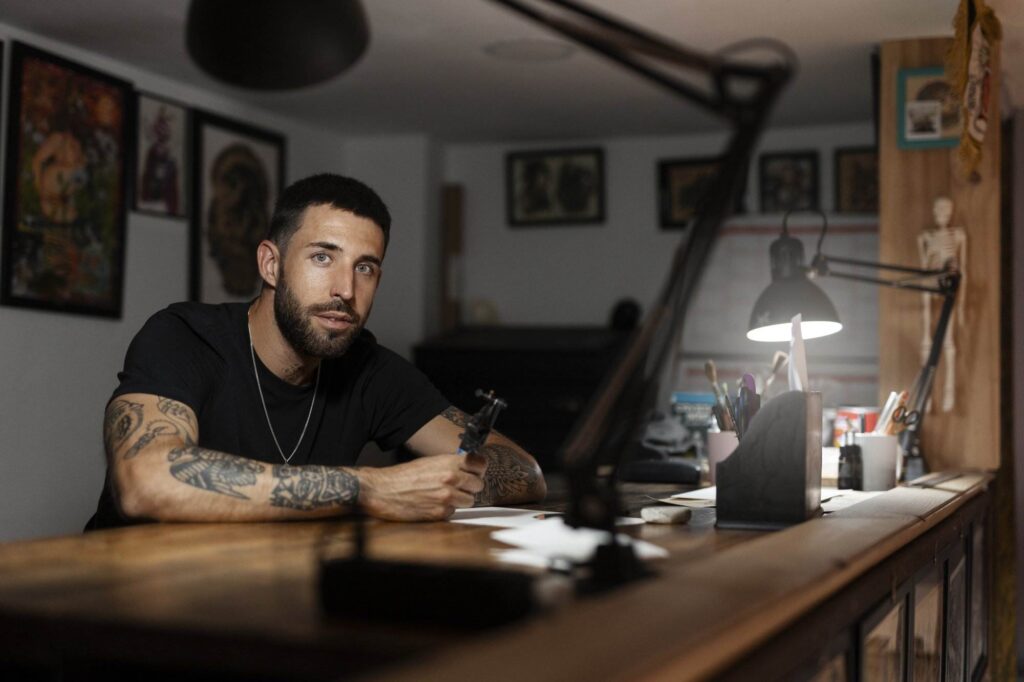 A man with tattoos sits at a dimly lit desk holding a tattoo machine in a workspace adorned with artwork and various desk items.