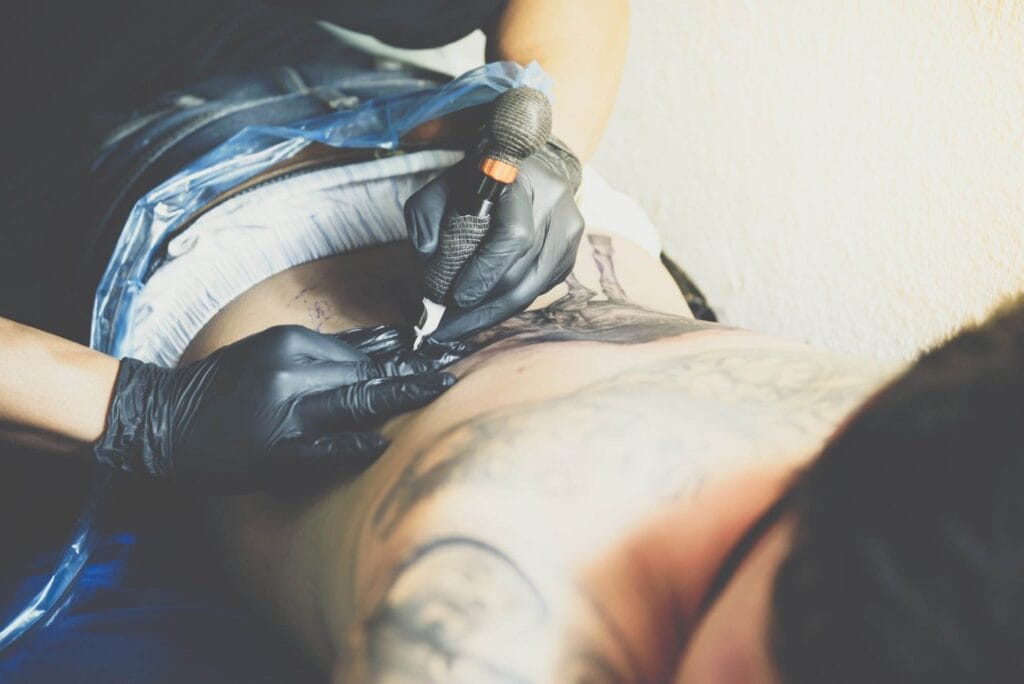 A tattoo artist, wearing black gloves, is applying a tattoo on a client's back using a tattoo machine. The client lies face down, partially covered by a white cloth.