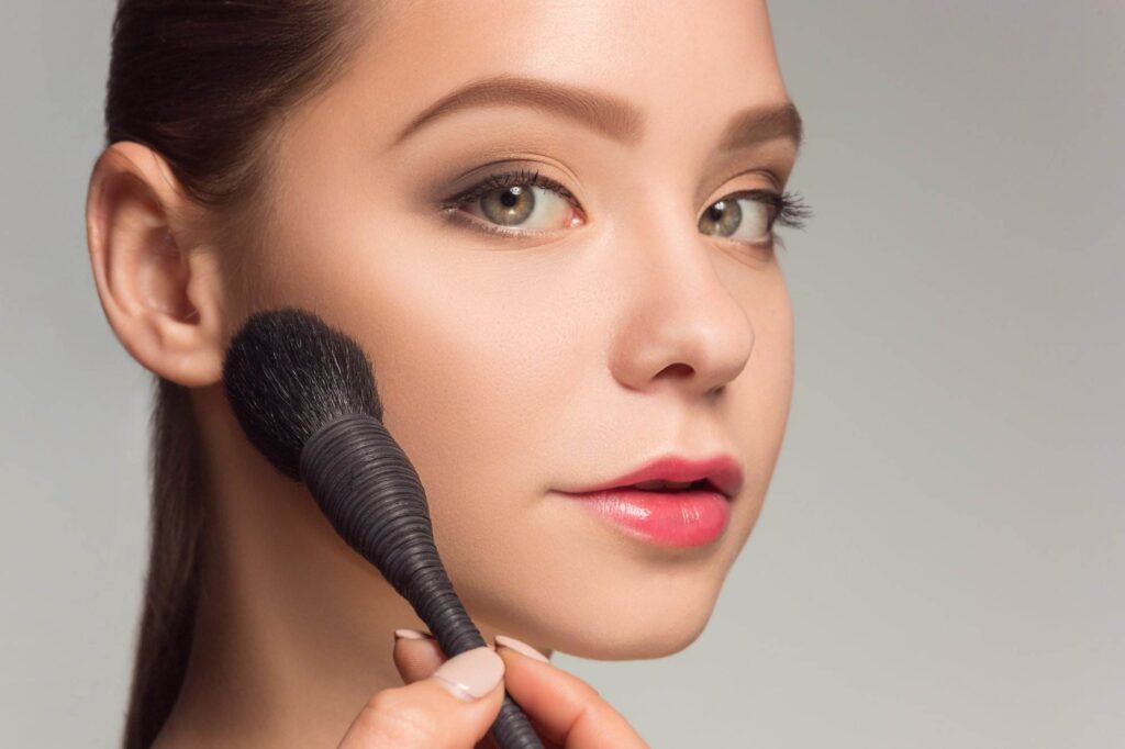 A person applies makeup with a brush to their cheek, focusing on cosmetic detail.