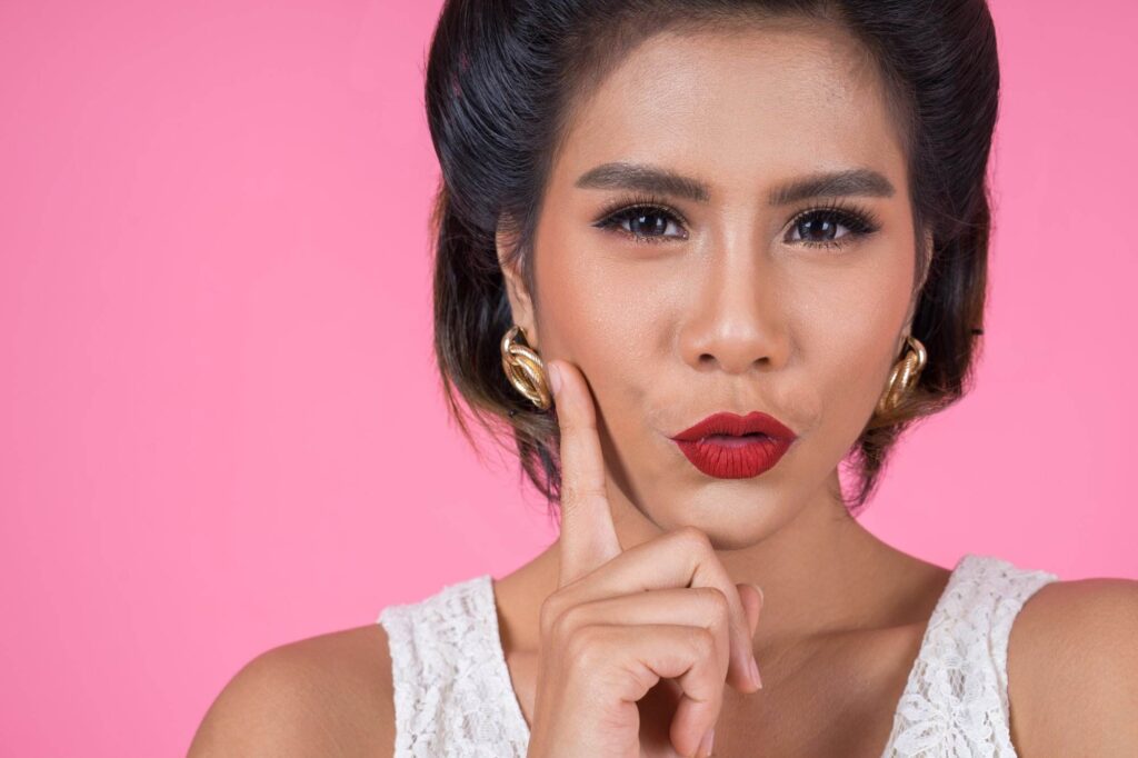 Woman with styled hair and red lipstick holding a finger to her cheek, against a pink background.