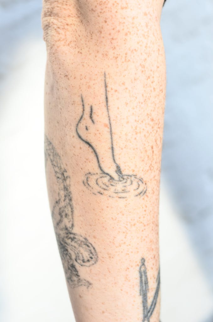 Crop unrecognizable person showing tattoos representing barefoot leg on tiptoe touching water on freckled skin