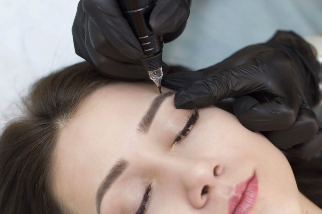 A person gets an eyebrow tattoo with a needle device. A technician wearing black gloves is performing the procedure.