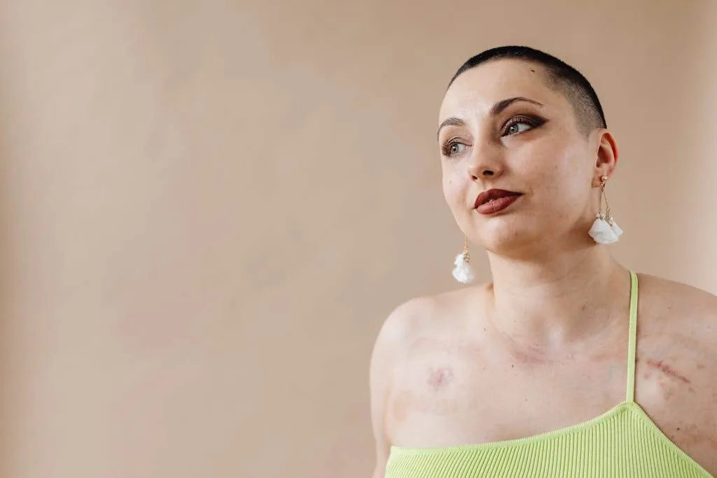 Woman with a shaved head wearing a lime green top and white floral earrings, looking to the side.