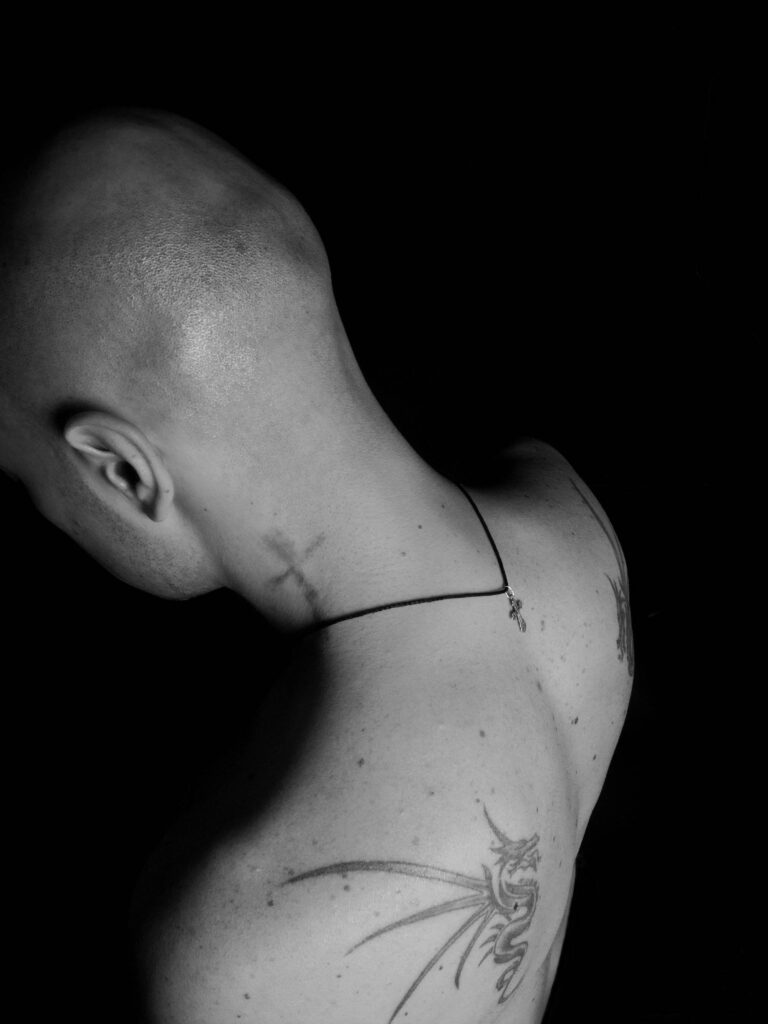 A black and white photo of a bald person facing away, showing tattoos of a cross on the neck and a dragon on the shoulder. A necklace with a small pendant is visible.