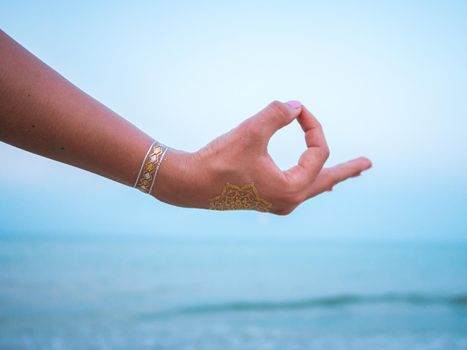 A hand with a henna design is shown in a yoga mudra gesture against a backdrop of a calm sea and blue sky.