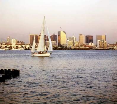 A sailboat on calm water with a city skyline in the background during sunset.