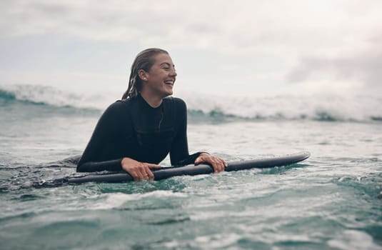 Person in a wetsuit lying on a surfboard in the ocean, smiling with waves in the background.