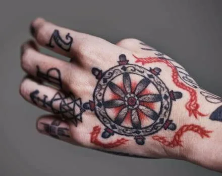 A hand tattoo featuring a detailed ship wheel with red accents surrounded by red dragons and other black ink symbols on the fingers and hand.