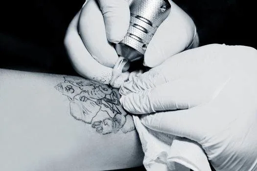 Close-up of a tattoo artist wearing gloves, using a tattoo machine to create a design on a person's arm, with a cloth wiping excess ink.
