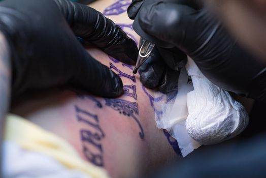 Close-up of a tattoo artist's gloved hands applying ink on a person's skin. The tattoo design includes intricate patterns and Roman numerals.