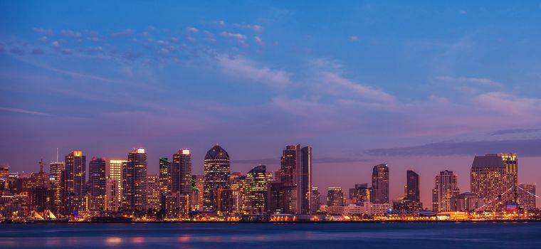 City skyline at dusk with illuminated buildings reflecting on calm water under a colorful sky.