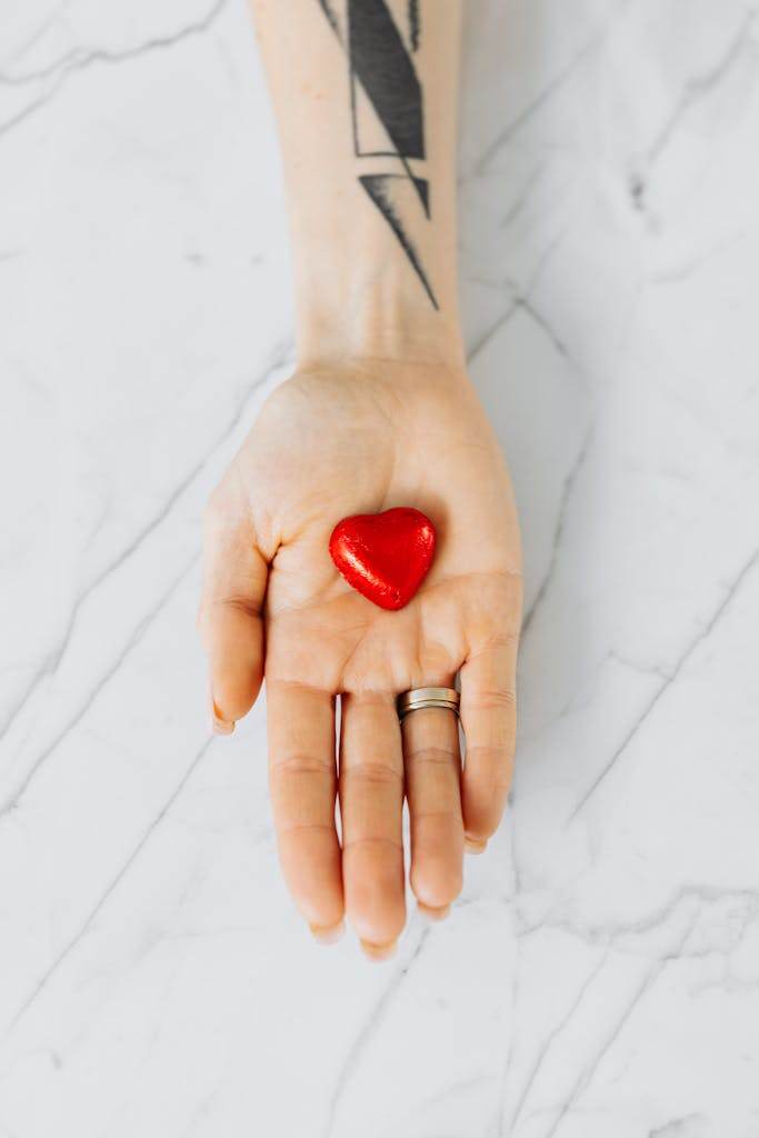 A tattooed hand holding a red heart-shaped object on a marble surface, symbolizing love and care.