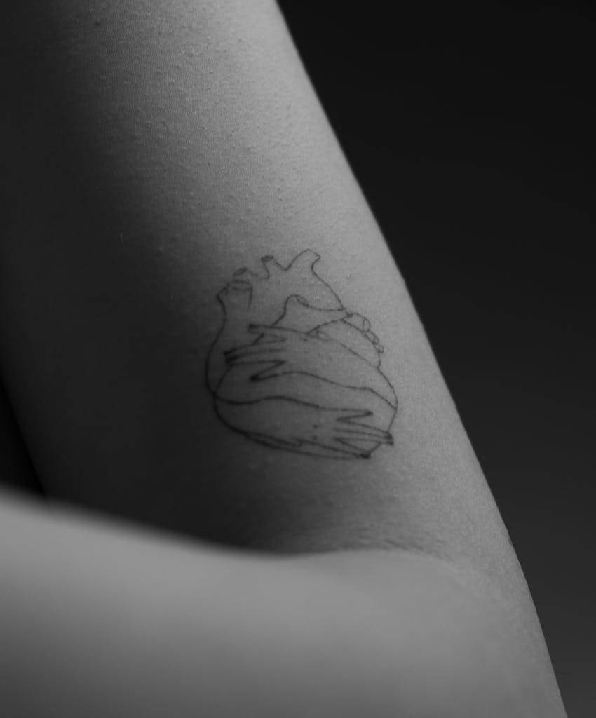 Black and white close-up of a heart tattoo on an arm, showcasing minimalist line art.