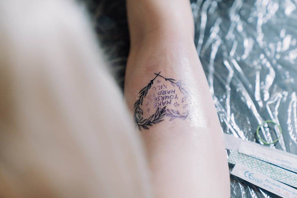 Close-up of a fresh tattoo with inspirational text encircled by laurel wreath on skin.