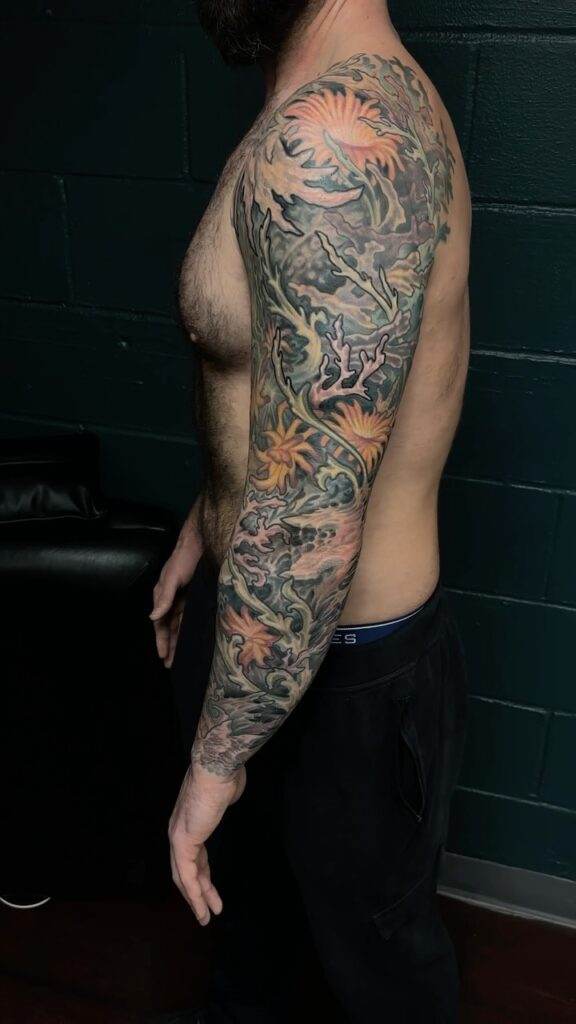 Man with a detailed full sleeve tattoo on his right arm, featuring vibrant colors and intricate designs, stands against a dark wall background.