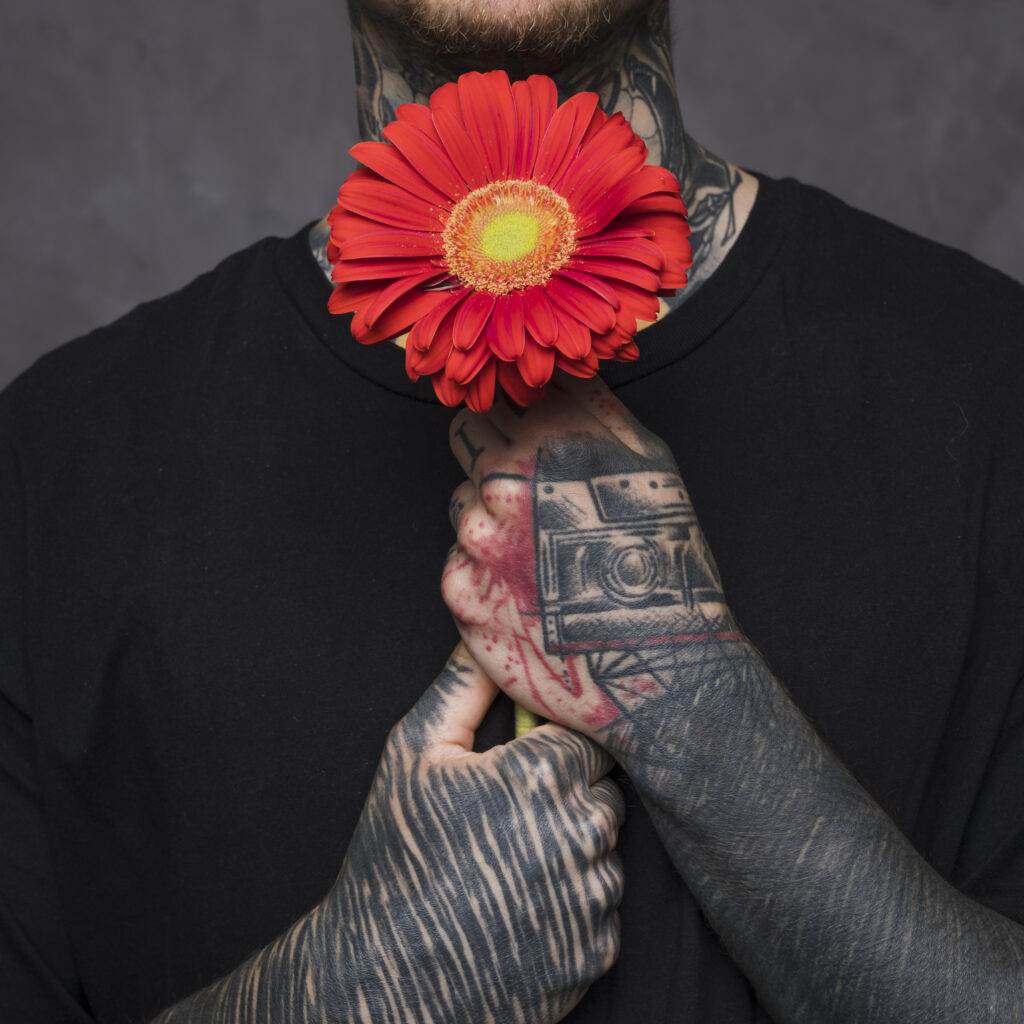Person with tattoos holding a red flower near their neck against a dark background.