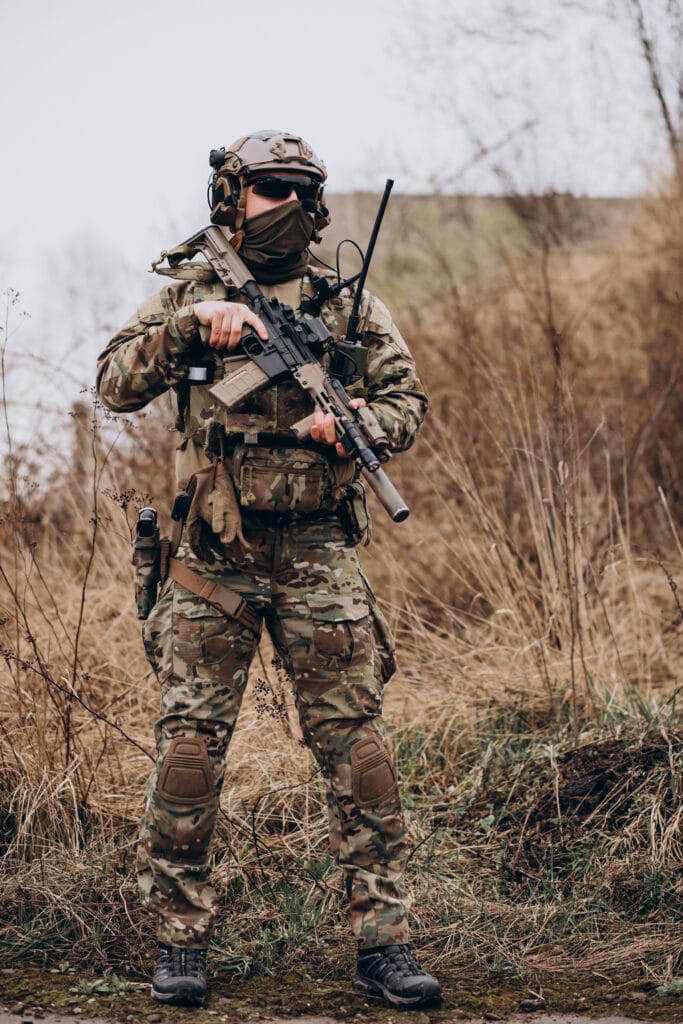 A soldier in camouflage uniform and tactical gear stands outdoors holding a rifle, with dry grass and overcast sky in the background.