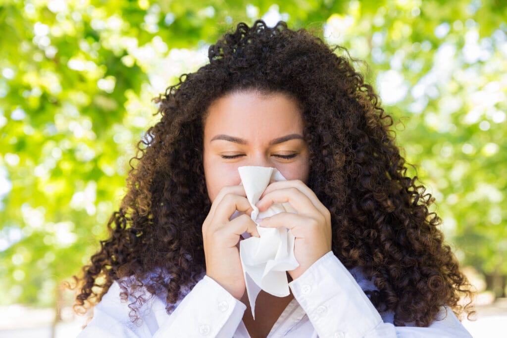 Woman with curly hair sneezing into a tissue outdoors on a sunny day.