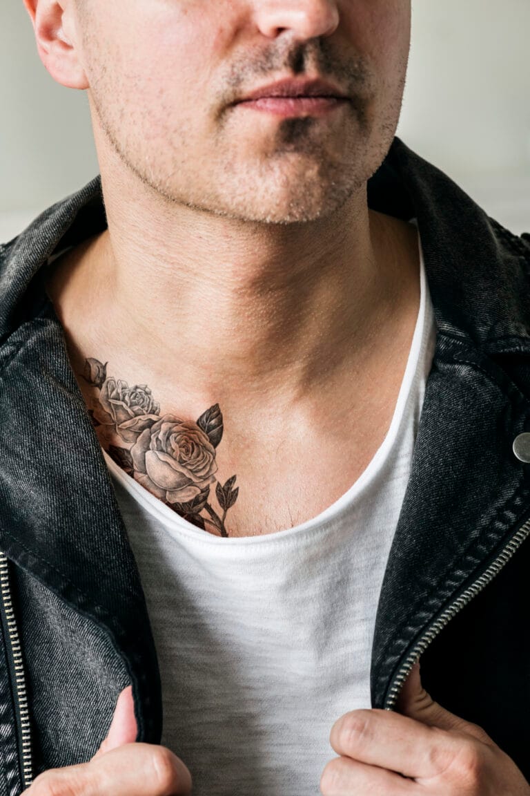 A man wearing a white shirt and black denim jacket shows a floral tattoo with roses on his left upper chest.