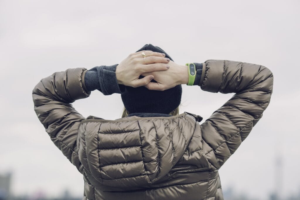 Person wearing a puffer jacket and a beanie stands outdoors with their hands clasped behind their head, facing away from the camera.