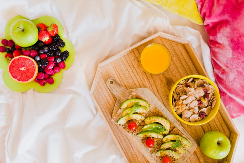 A breakfast spread on a bed with avocado toast, orange juice, cereal, an apple, and a plate of assorted fruits including grapefruit, berries, and apples.