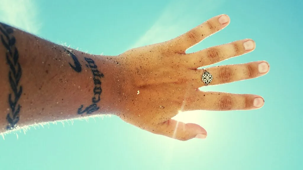 Hand with a ring, tattoos, and sand on fingers, reaching toward a bright blue sky.