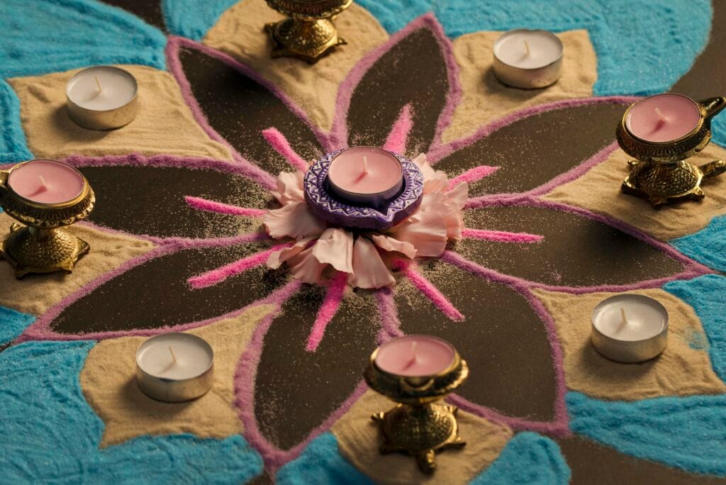 A colorful rangoli design with tealights and candle holders arranged in a circle on a floor, featuring shades of blue, purple, beige, and pink.