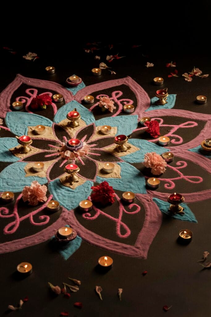 A colorful rangoli design with pink, blue, and black patterns is decorated with candles, oil lamps, and flowers on a dark surface.