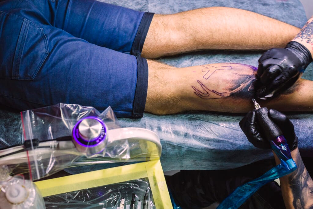A tattoo artist in black gloves works on a man's lower leg, following a purple stencil outline above the knee while the man lies on a padded table.