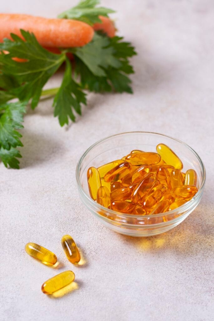 Softgel capsules in a glass bowl with a carrot and leafy greens in the background.