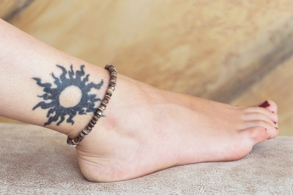 A close-up of a person’s ankle with a black sun tattoo and a beaded anklet, against a neutral, blurred background.