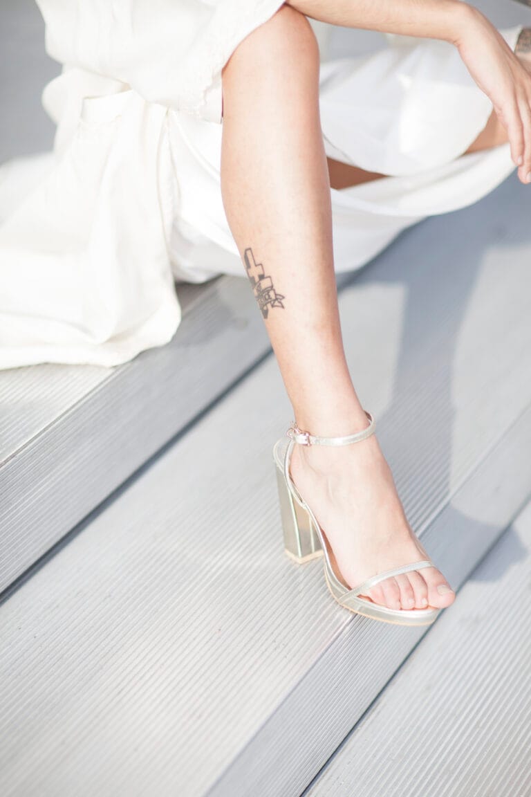 A person in a white outfit sits on gray steps, showing a tattoo on their calf and wearing high-heeled, open-toe sandals.