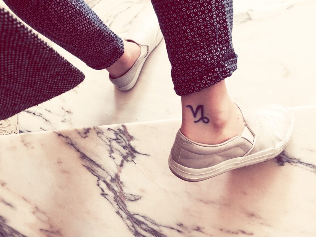 A person wearing patterned pants and white slip-on shoes with a Capricorn zodiac symbol tattoo on their ankle, standing on a marble surface.