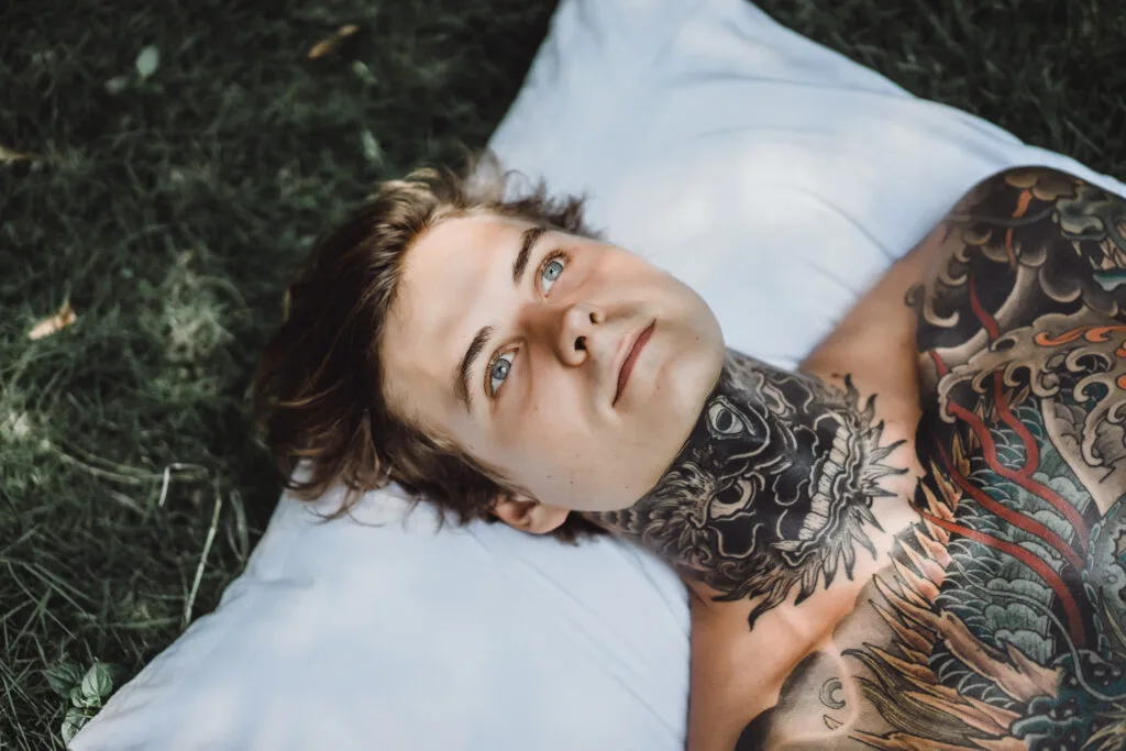 A young person with blue eyes and extensive neck and chest tattoos lies on a white pillow outdoors, looking upward.