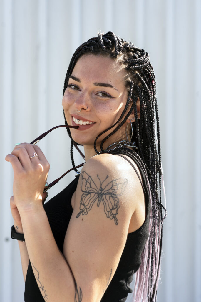 A woman with braided hair, a butterfly tattoo on her upper arm, and a nose ring smiles while holding sunglasses near her mouth.