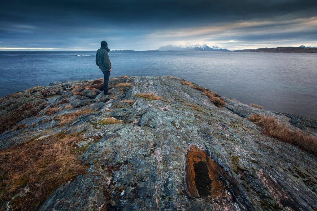 Person standing on rocky shoreline overlooking a calm ocean under a cloudy sky.