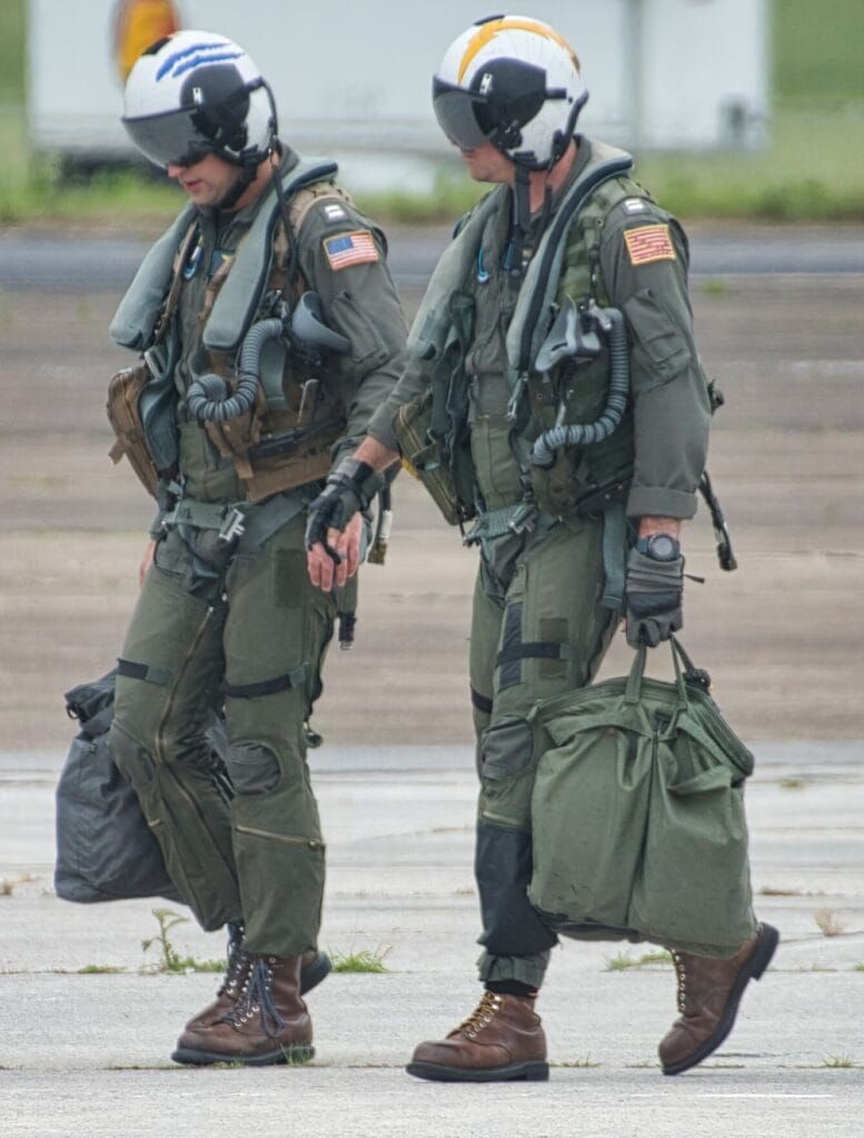Two pilots in flight suits and helmets walk side by side on a runway, each carrying a large duffel bag.
