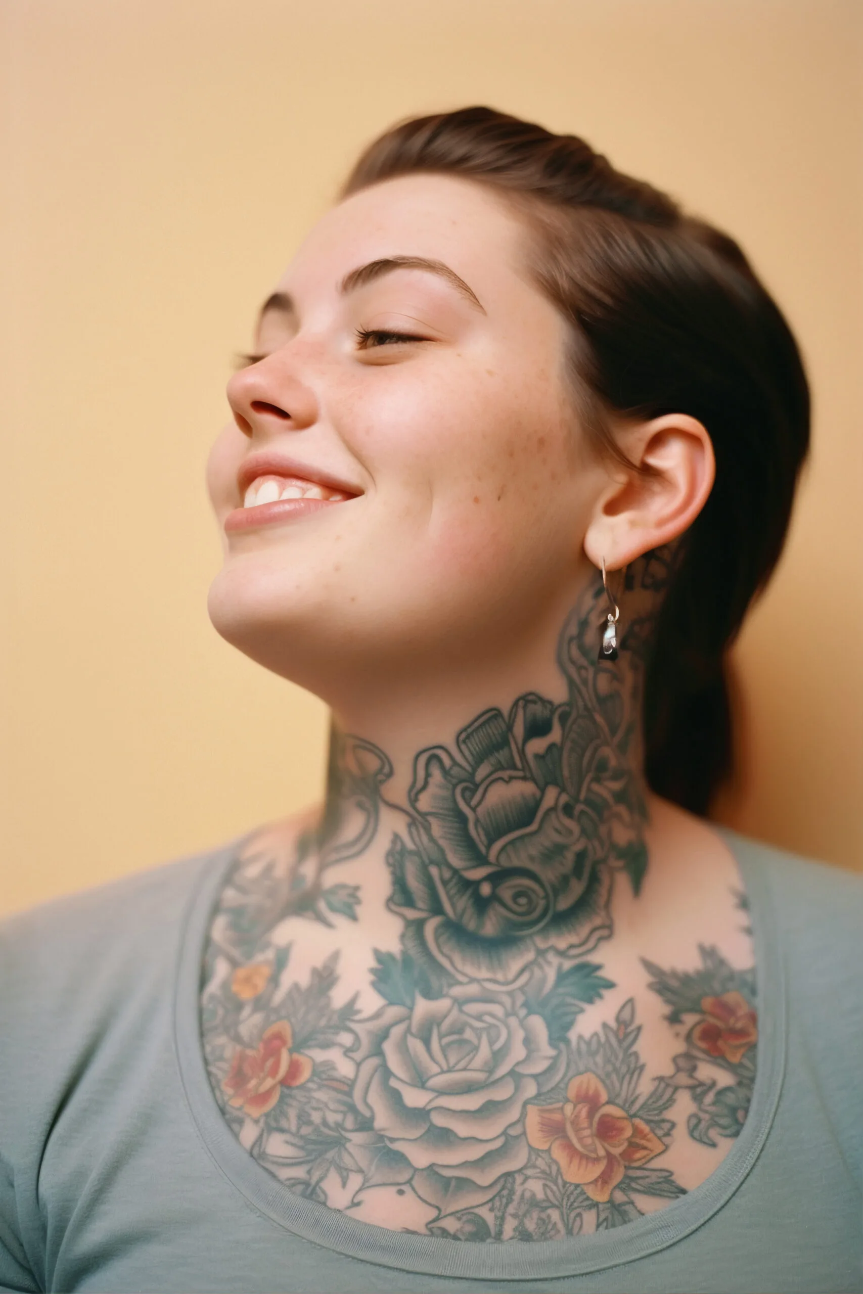 A young woman with short dark hair smiles to the side, showing detailed floral tattoos on her neck and upper chest. She wears a light green top and a small hoop earring.