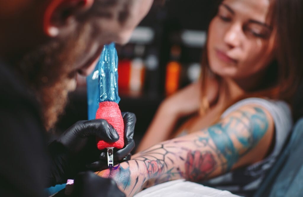 Tattoo artist wearing black gloves, holding a tattoo machine, working on a womans colorful arm tattoo.