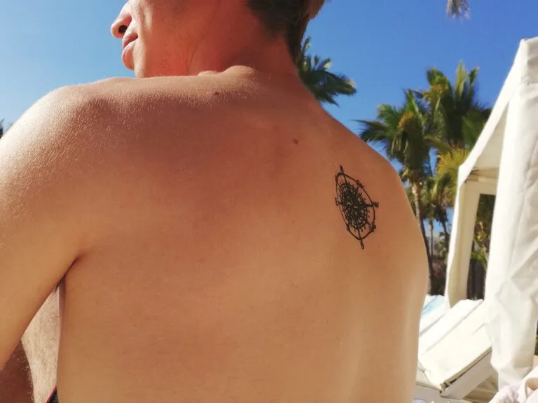 A person with short hair sits outdoors, displaying a black compass tattoo on their upper back. Palm trees and a white structure are visible in the background under a clear sky.