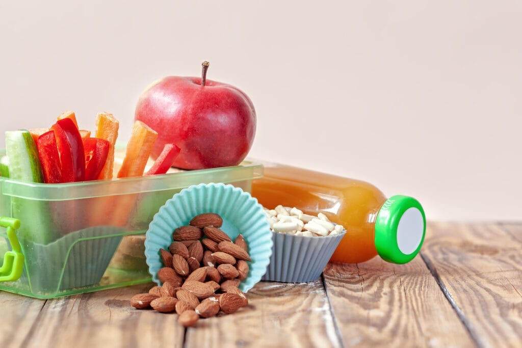 Red apple, carrot sticks, almonds, white snack, and honey bottle on a wooden surface.