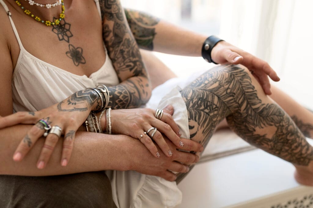 Two people with multiple tattoos and jewelry sit close together; one has their hand resting on the other's leg, both wearing light clothing.