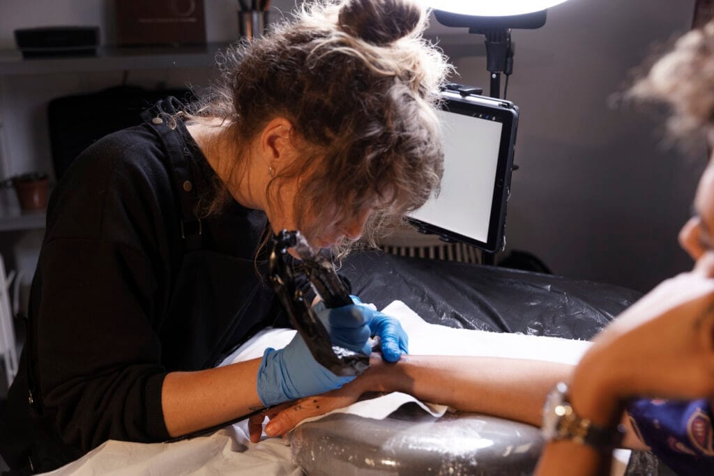 Tattoo artist in black shirt and blue gloves tattooing a persons forearm.