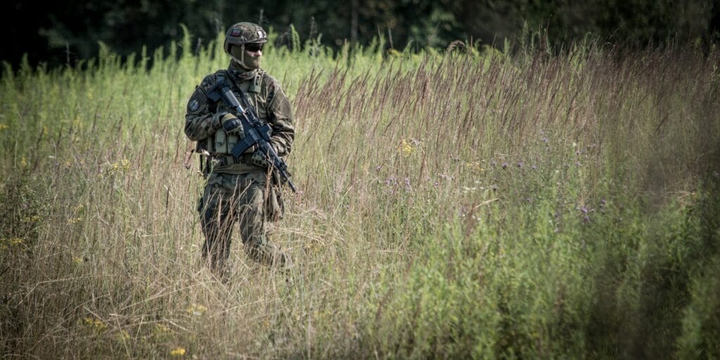 A person in military gear and helmet holding a rifle stands in tall grass and wild plants outdoors.