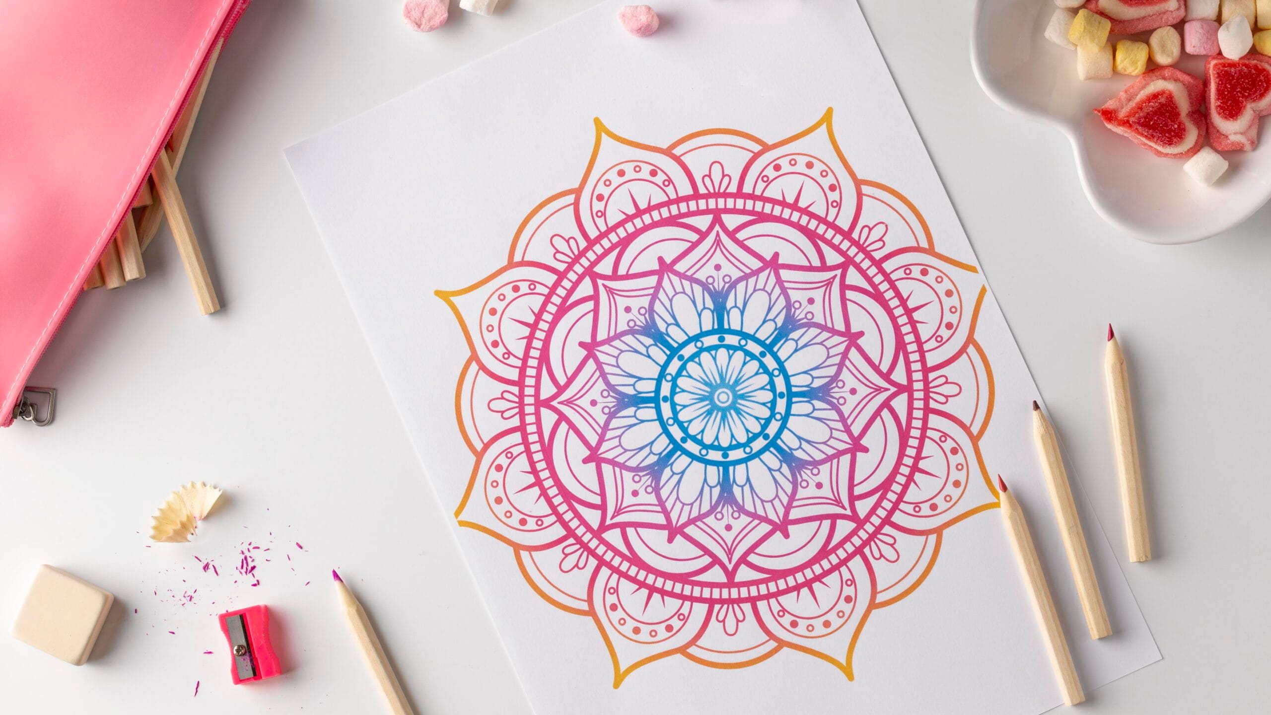 A colorful mandala drawing on white paper, surrounded by colored pencils, erasers, pencil shavings, and a tray with dried fruit snacks on a white surface.